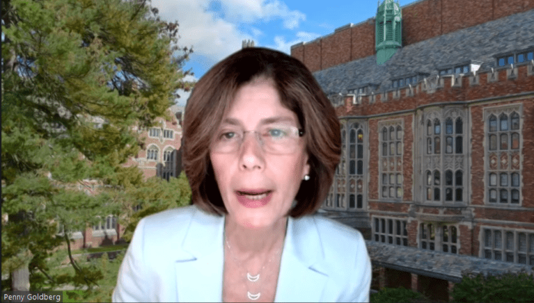 Penny Goldberg on Zoom with a virtual background of trees and a brick building.