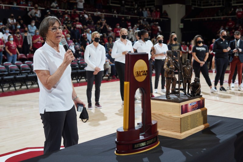 Stanford women's basketball head coach Tara VanDerveer speaks into a microphone with the NCAA Basketball championship on a table in front of her.