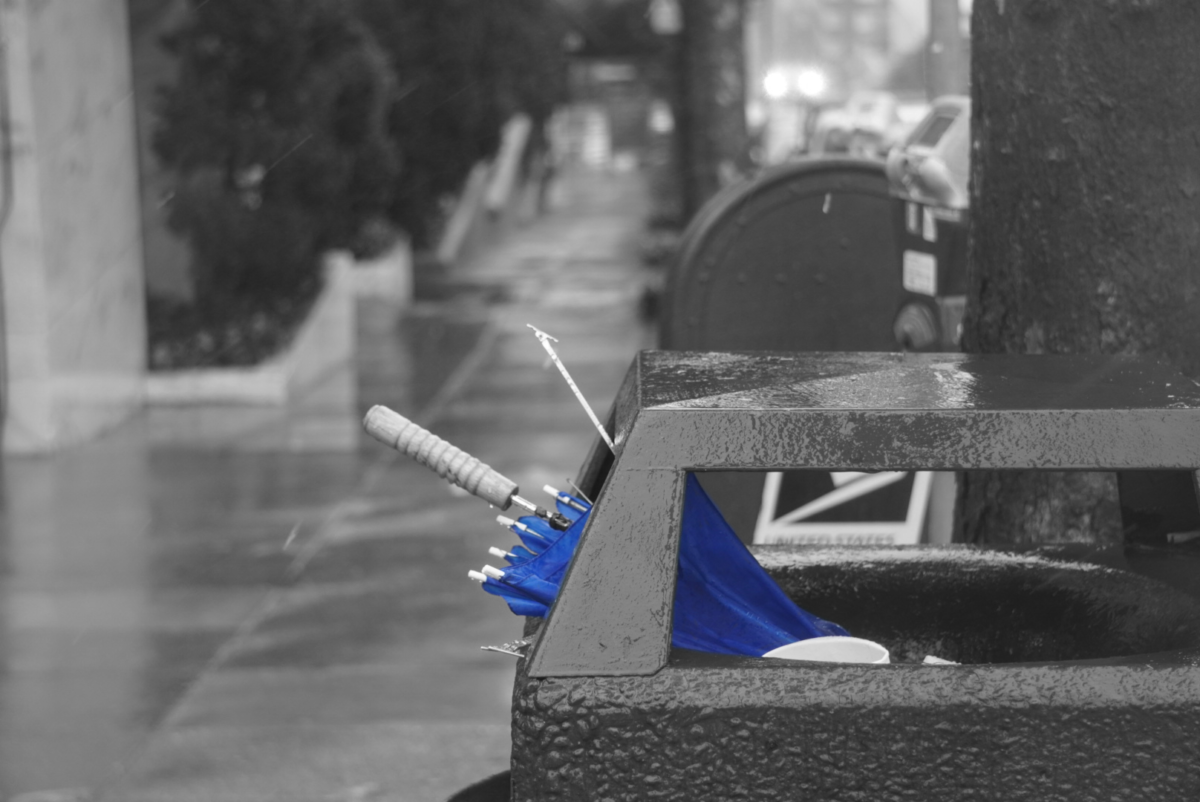 An umbrella, broken by the strong winds, sits in a trash can. The umbrellas shines blue while everything else is picture grayscale.