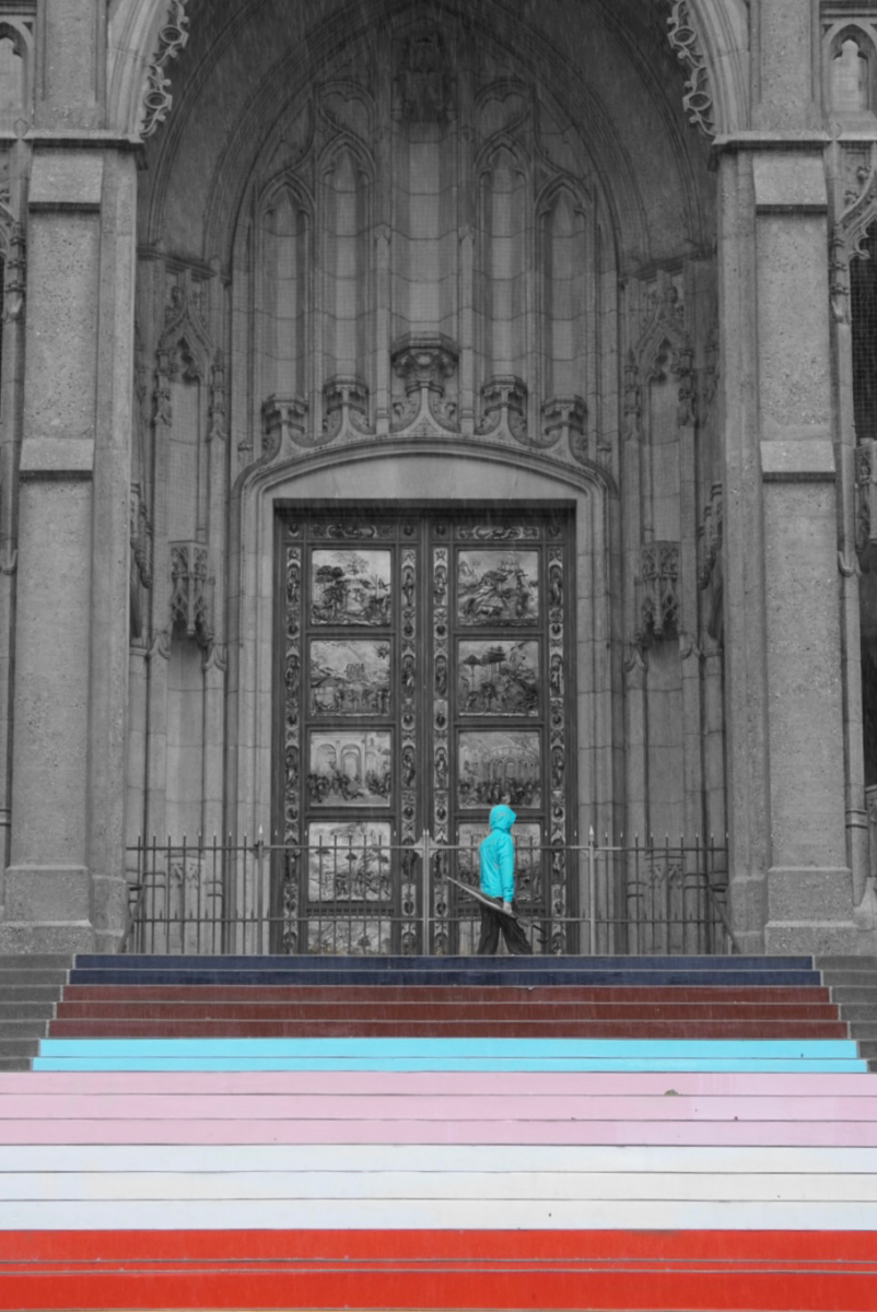 A woman stands in front of Grace Cathedral after walking up the Pride Month steps. The steps and the woman's jacket are colorful while the church is pictured grayscale.