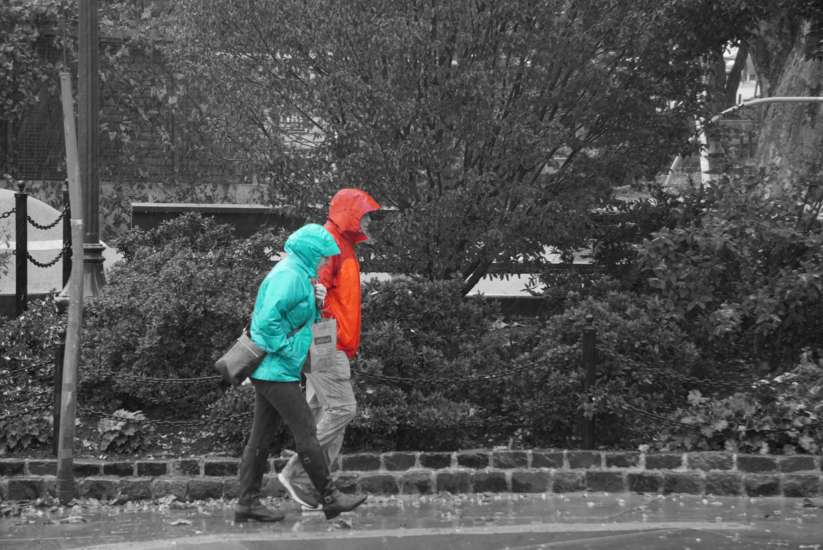 A couple walks through the rain together. Their jackets shine red and turquoise while everything else is pictured grayscale. 