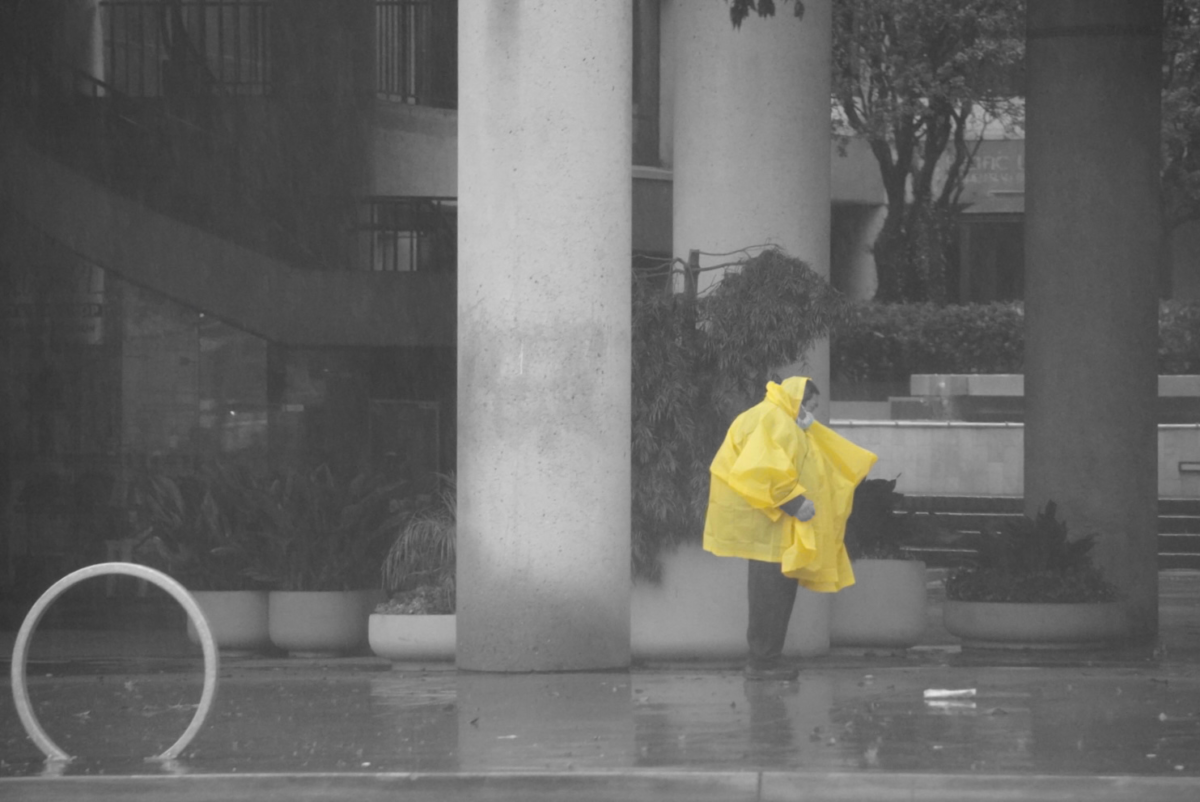 A man attempts to shield himself from the downpour with his yellow poncho. The poncho shines yellow while everything else is pictured grayscale.