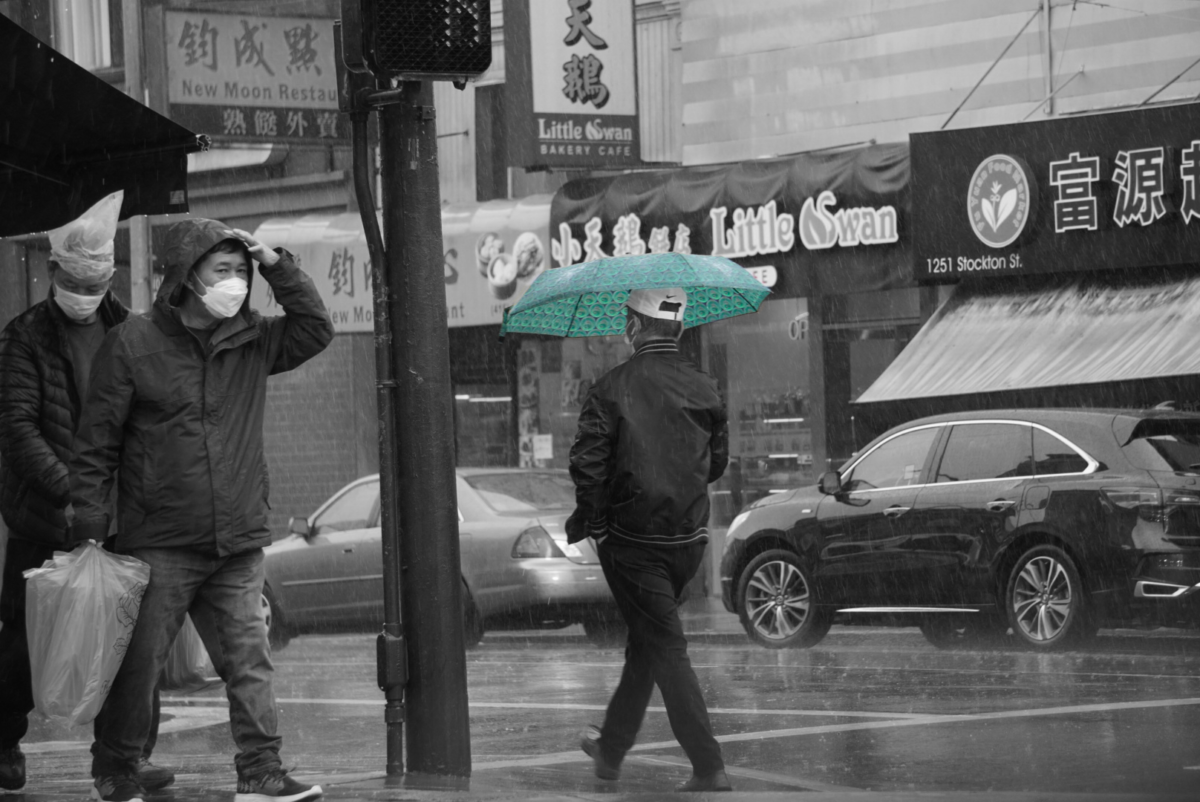 A man walks through ChinaTown streets, protecting himself from the rain with an umbrella. The umbrellas shines green while everything else is pictured grayscale.