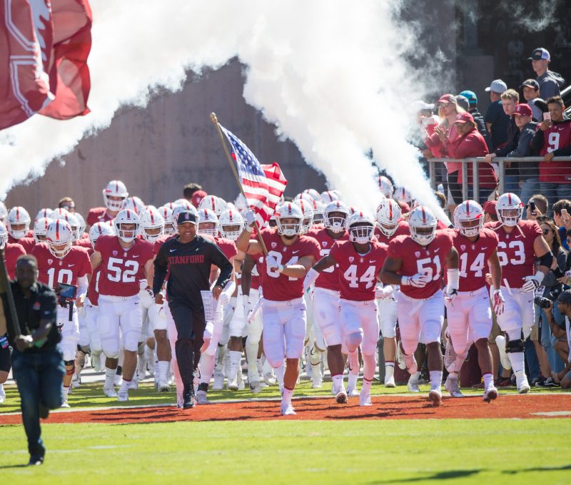 A group of Stanford football players runs out of the tunnel, with one carrying an American flag, and fake smoke in the air behind the players.