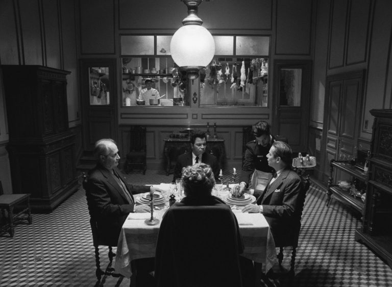 A black and white photo of four people sitting around a table with a waiter serving them and a cook looking on from afar in the kitchen