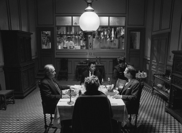 A black and white photo of four people sitting around a table with a waiter serving them and a cook looking on from afar in the kitchen