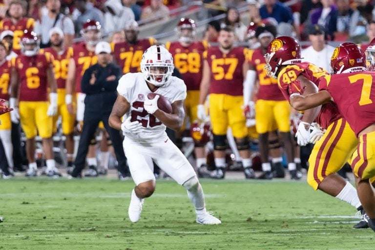 A running back runs up the field carrying the football with several playing running at him from the front and several players in the background on the sidelines.