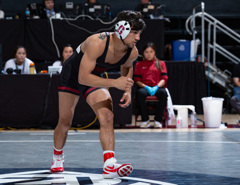 A wrestler gets in his ready stance on a wrestling mat with several people in the background watching from the sidelines.