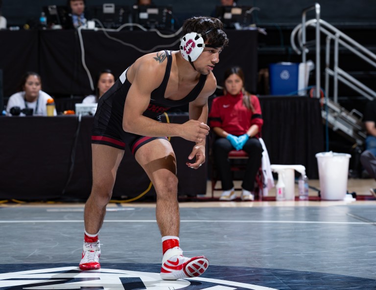 Real Woods gets in his ready stance on a wrestling mat with several people in the background watching from the sidelines.