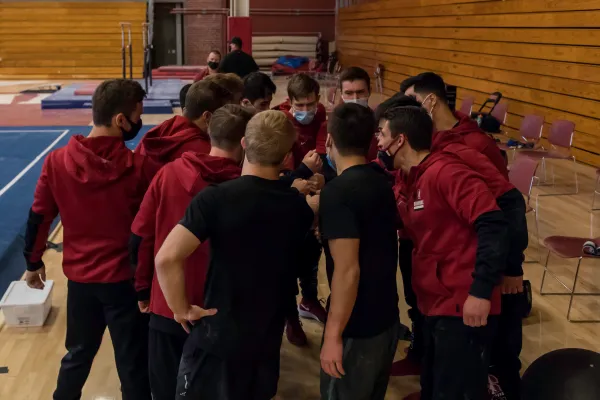 A group of men's gymnastics athletes huddles together for a talk during a meet.