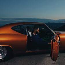 Singer-songwriter Khalid sits inside an orange car with the door open with a darkened sky as the backdrop.