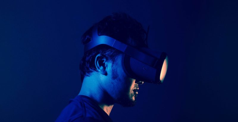 A man wears a virtual reality headset, facing to the right under blue light.