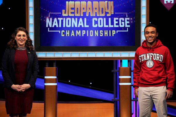 Issac Applebaum is shown in a Stanford sweatshirt, standing in front of a blue screen that says "Jeopardy! National College Championships"