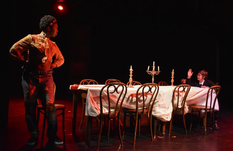 Figure in red light stands at the head of candle lit table with another person seated at the end.