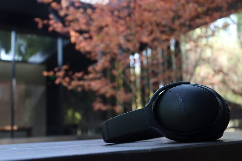 A pair of Bose headphones rest on a black surface in front of an out-of-focus red-leaved tree and glass building.