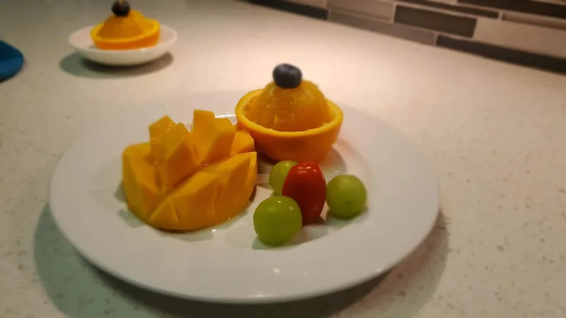 A plate of assorted fruits, including 3 grapes, a spliced mango, and an orange resting in half of the peel, topped with a blueberry.
