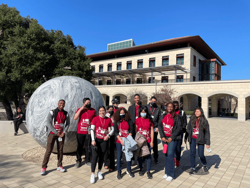 Students in red SERIS shirts posing in front of a brick building and large ball sculpture.