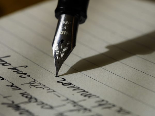 Close shot of a fountain pen and paper; the pen is in the middle of writing something in neat cursive penmanship.