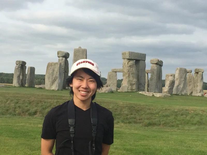 Dylan Simmons stands smiling in front of Stonehenge with a camera.