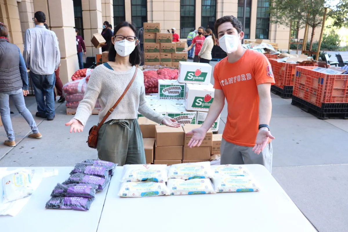 Two student volunteers pose, smiling, with arms spread wide behind table with rice and bean packets. Boxes of produce behind them. 