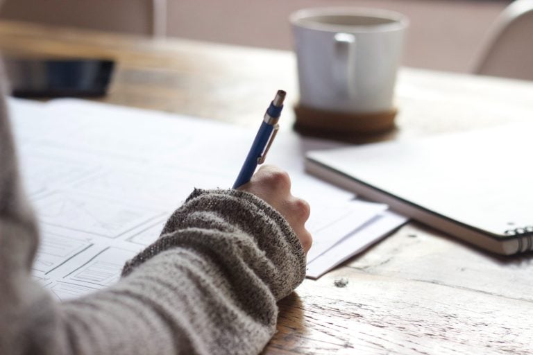 A hand writes on a piece of paper on a desk with a cup of coffee in the background.