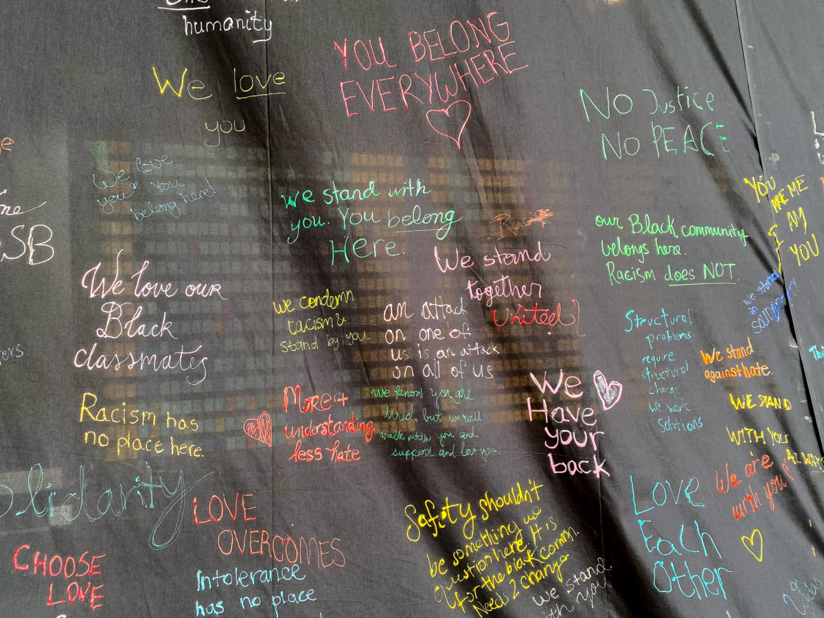 A black cloth hangs across a building wall with handwritten messages of solidarity in multiple colors. 