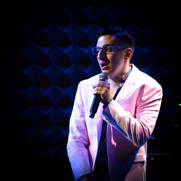 Photo of Ryan standing under the spotlight against a neon blue backdrop and holding a microphone. Ryan is wearing neon blue glasses, a pink blazer and a floral collared shirt underneath.