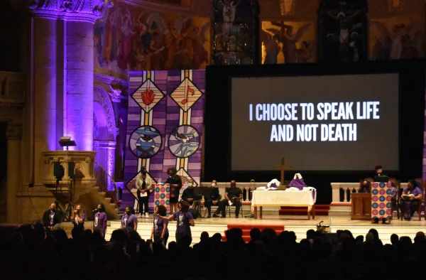 Several people sit on a stage inside Memorial Church. A projected screen reads "I choose to speak life and not death." Several singers are at microphones at the front.