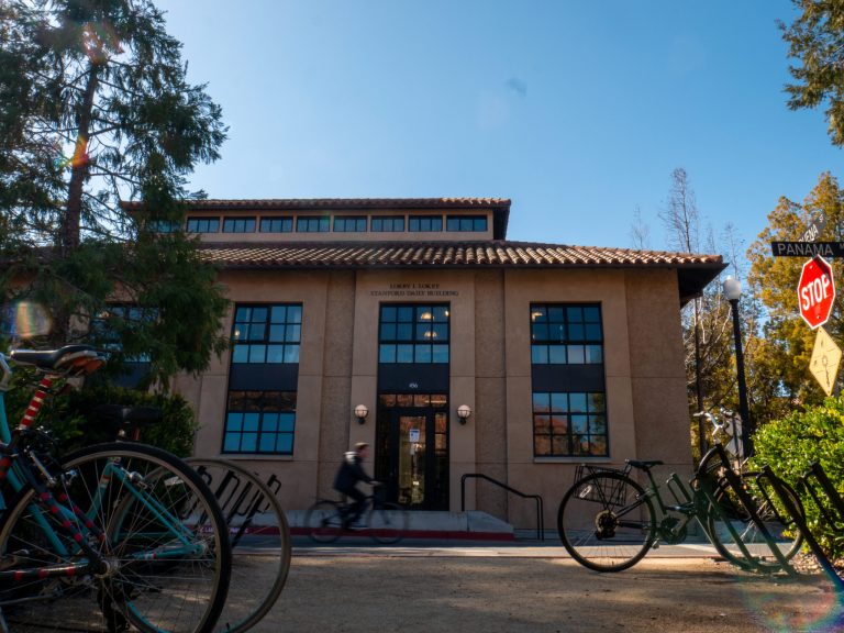 Picture of Lorry I. Lokey Stanford Daily Building with bike racks and bikes at front of image, man biking by