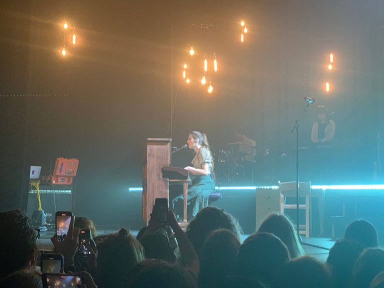 An array of old lightbulbs hangs from the ceiling, glowing orange. Dodie sits at the piano, singing for hundreds of fans in front of a streak of lightning blue LED lights.