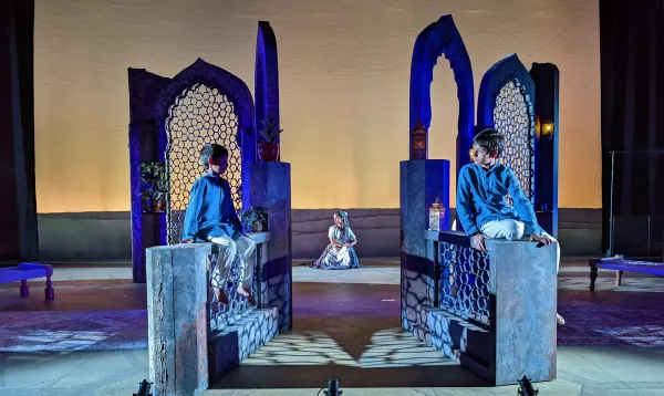 The two young actors playing the role of "Pali" sat on the two fences on each side of the stage facing each other. A lady in white robe with her hair tied to one side sat on the ground mid-dancing, at center-back stage, behind the two boys and the set.