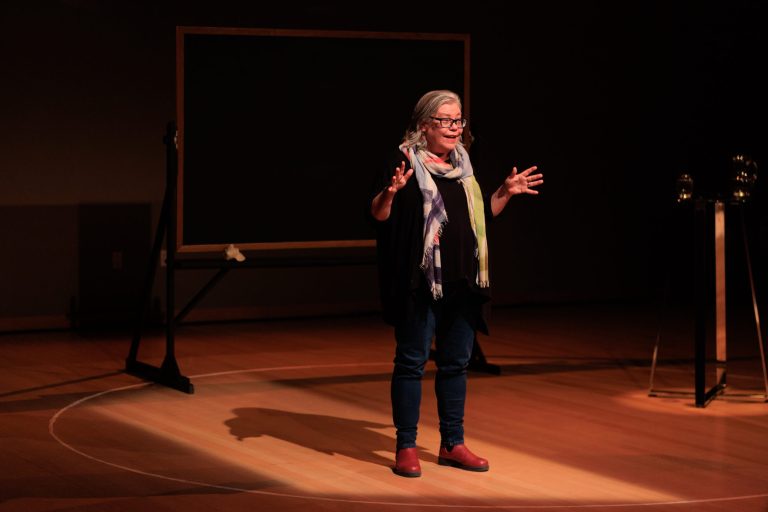 Alanna Mitchell speaks to the audience with both hands up, spotlighted in front of a blackboard