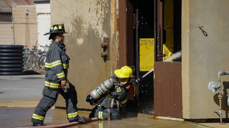 Two firefighters. One sprays water into a building while the other walks nearby.