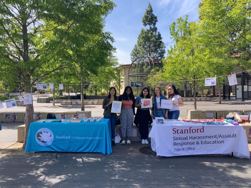 Five female-identifying students stand by two tables in White Plaza, promoting sexual violence prevention and support resources as part of Denim Day.