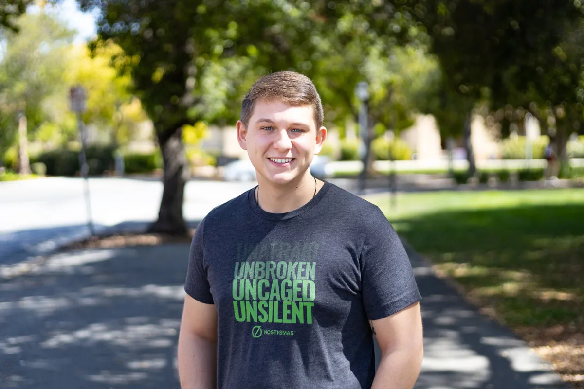 Conner Oberhauser smiles for a photo in front of trees. His tshirt reads "Unafraid,  Unbroken, Uncaged, Unsilent. NoStigmas"