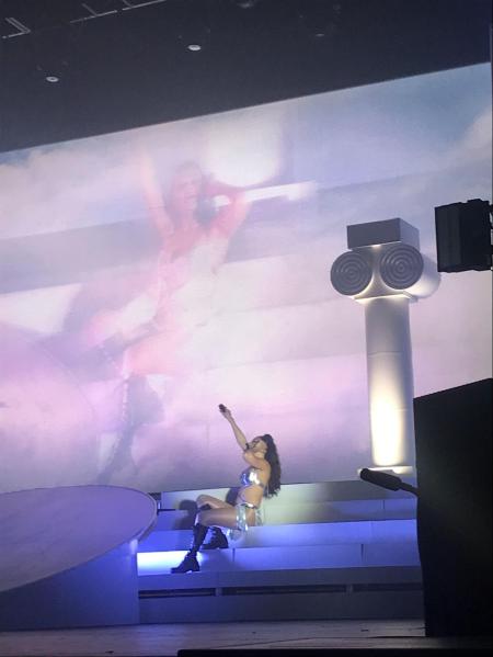 Charli XCX, wearing a silver bikini and boots, sits on the steps of her Fox Theater set while holding a microphone up. Behind her is a large close-up projection of her onstage