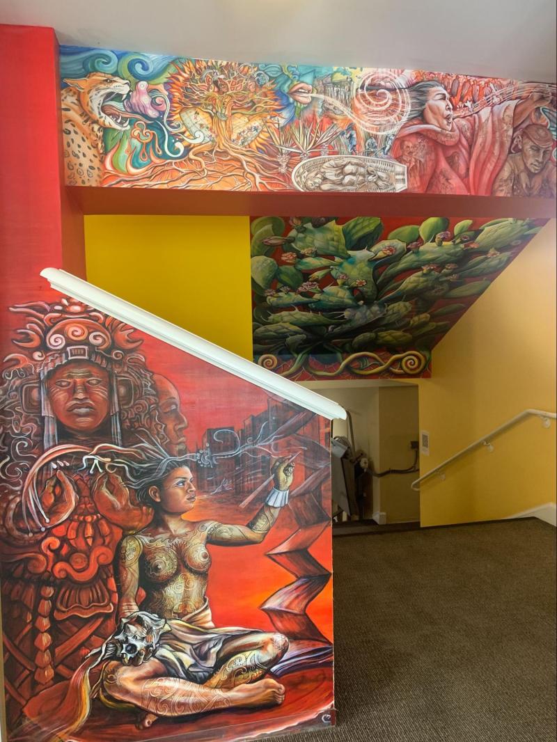 The mural in El Centro Chicano y Latino by Alicia. It features a shirtless Yucatán woman writing, scenes of genesis and slavery, and a nopal.