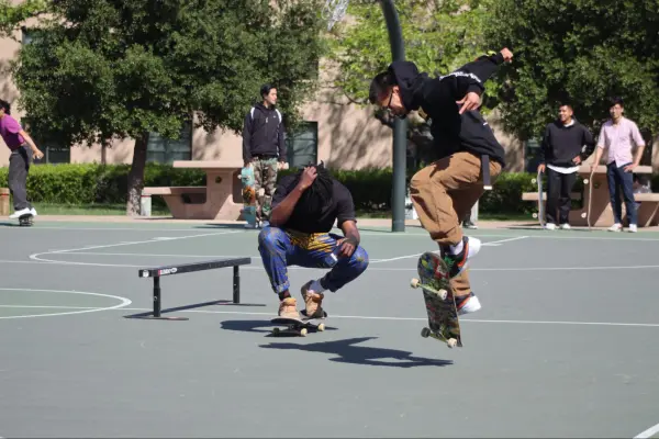 One skater hangs in midair, performing a flip trick, while another crouches down on their board, filming. Onlooking students watch from the side of the basketball court.