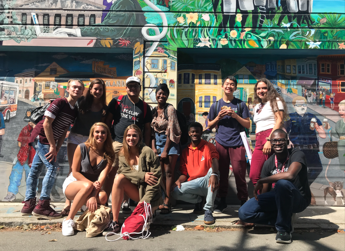 A group of 10 students pose and smile in front of a street mural. 