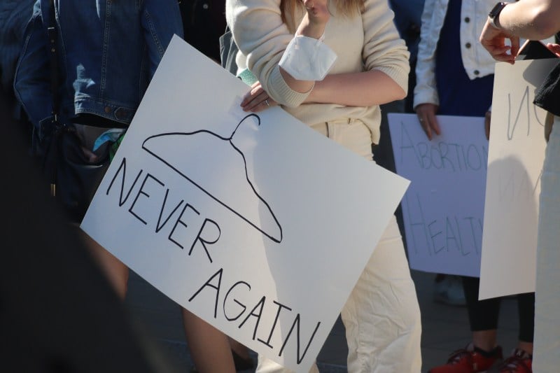 An unidentified female student holds a sign that has a photo of a coat hanger and says "never again." 