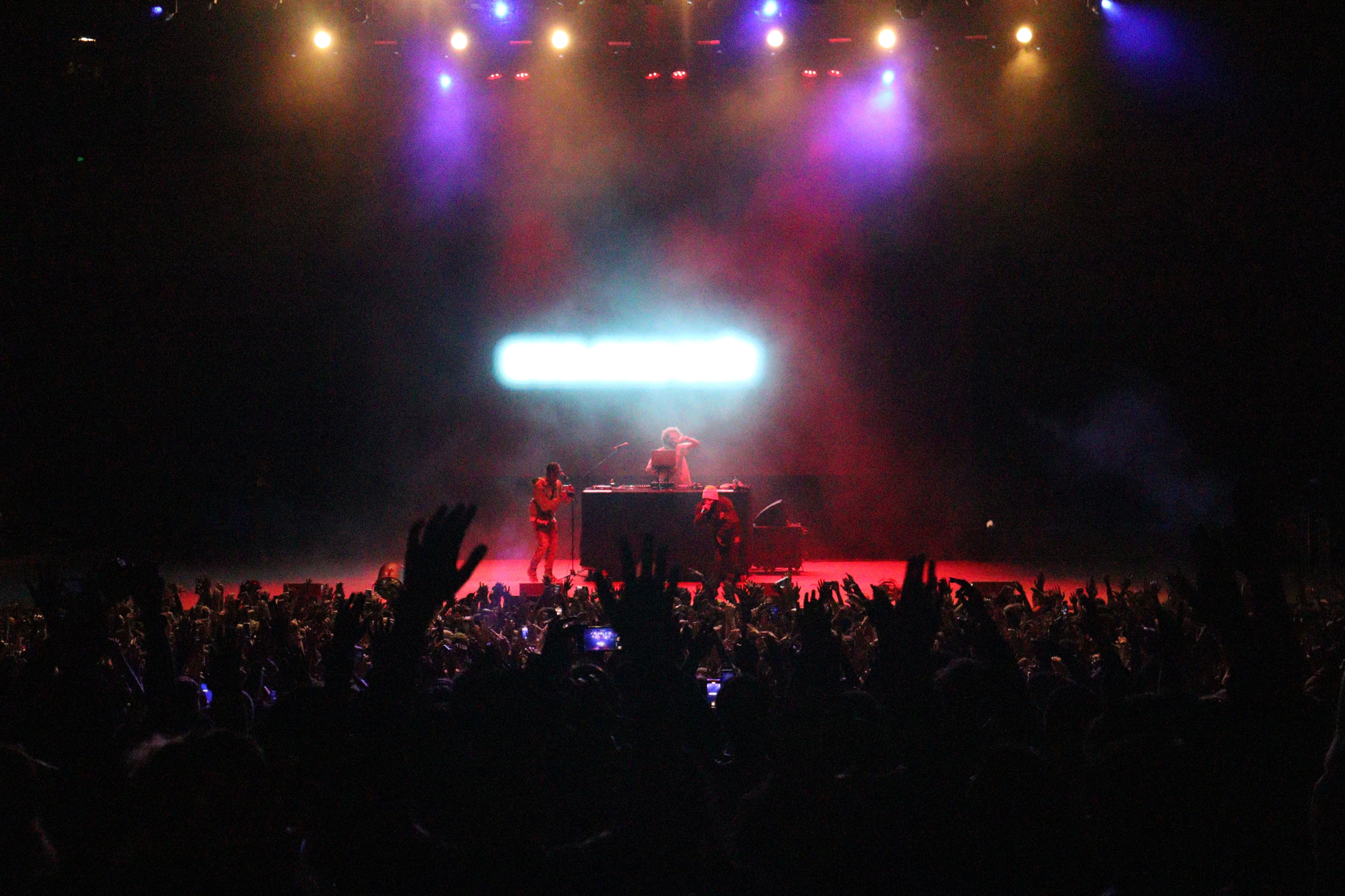 wide picture of three people performing on a stage, dark crowd on bottom half of image, orange and purple lights on the stage