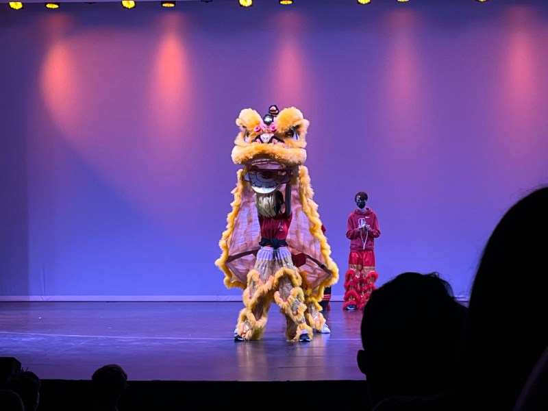 A life-sized yellow lion puppet operated by two students stands onstage. The front student lifts the front of the lion above her head as another student plays cymbals in the background.