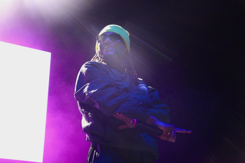 A black man stands in front of a bright purple fluorescent sign, wearing a dark green jumpsuit, a lime green beanie and sunglasses. He holds a microphone.