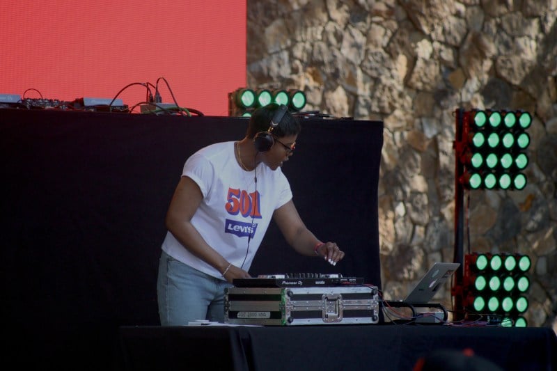 Student DJ Arielle Williams '22 stands on stage in front of a table and a DJ mixer. She is wearing headphones and a white t-shirt. She looks at her computer while touching the board.