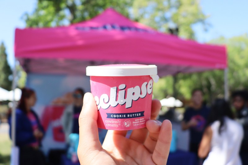 Hand holding up a paper cup of ice cream in front of a brightly coloured pink tent booth. The cup wrote 'eclipse' and below it 'cookie butter'