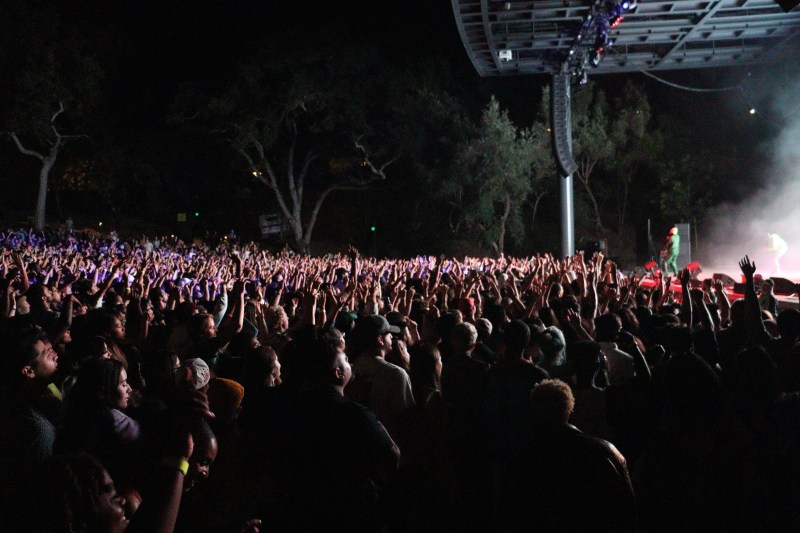wide shot of thousands of concertgoers raising arms up, watching amine on stage to the right.
