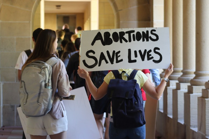 A group of students march near Green Library, with one student holding up a sign that says "abortions save lives"