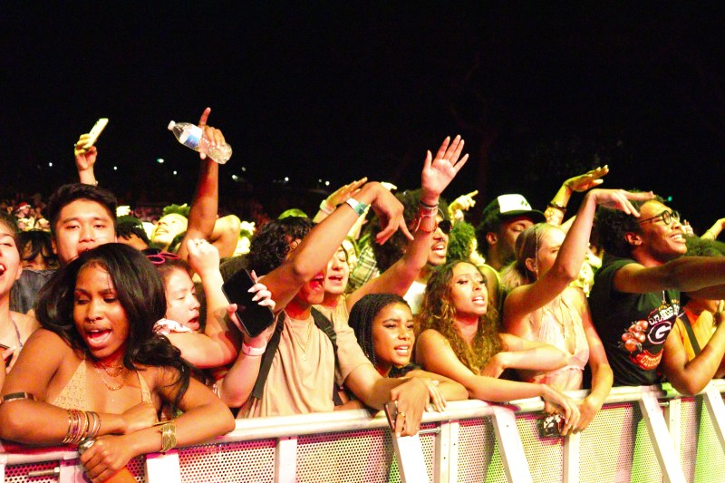 Dozens of students stand behind a barrier, very close together. Many point and gesture with their hands to the music, while some reach over the barrier toward the stage.