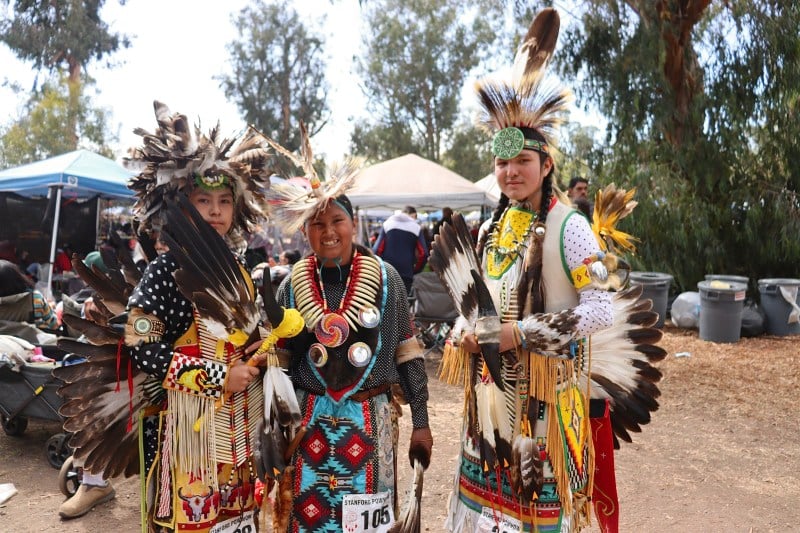 Three traditional dancers wearing regalia stand together for a photo.