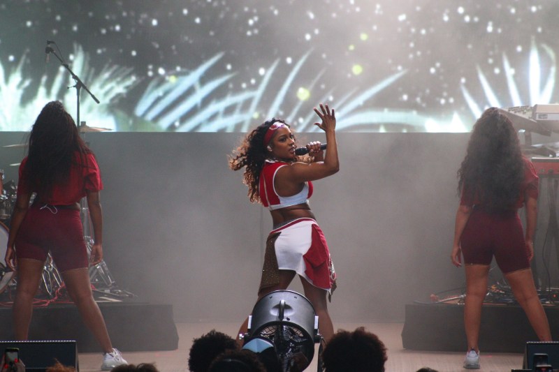 Three black women stand facing away from the camera, posing with their hips to one side. Victoria Monét, in the center, has her head turned toward the audience and a microphone to her mouth. The three wear custom Stanford cheerleading outfits.
